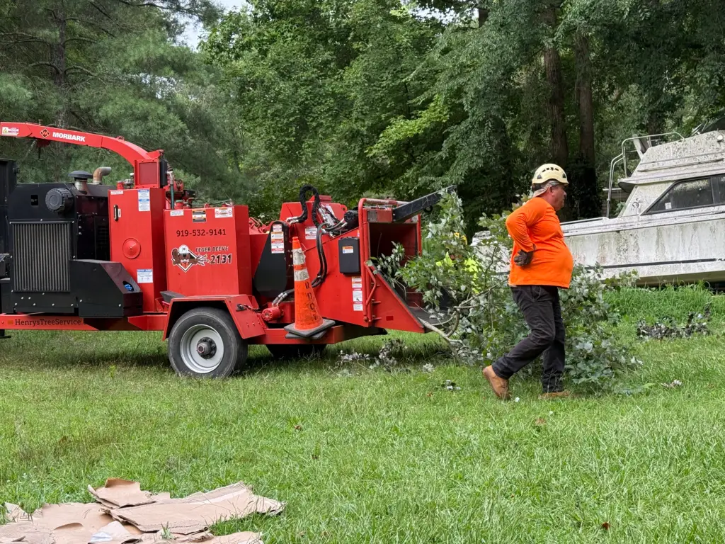 Tree service professional in orange shirt operating a red stump grinder, clearing branches in a grassy yard, with a boat in the background, showcasing Henry's Tree Service equipment.