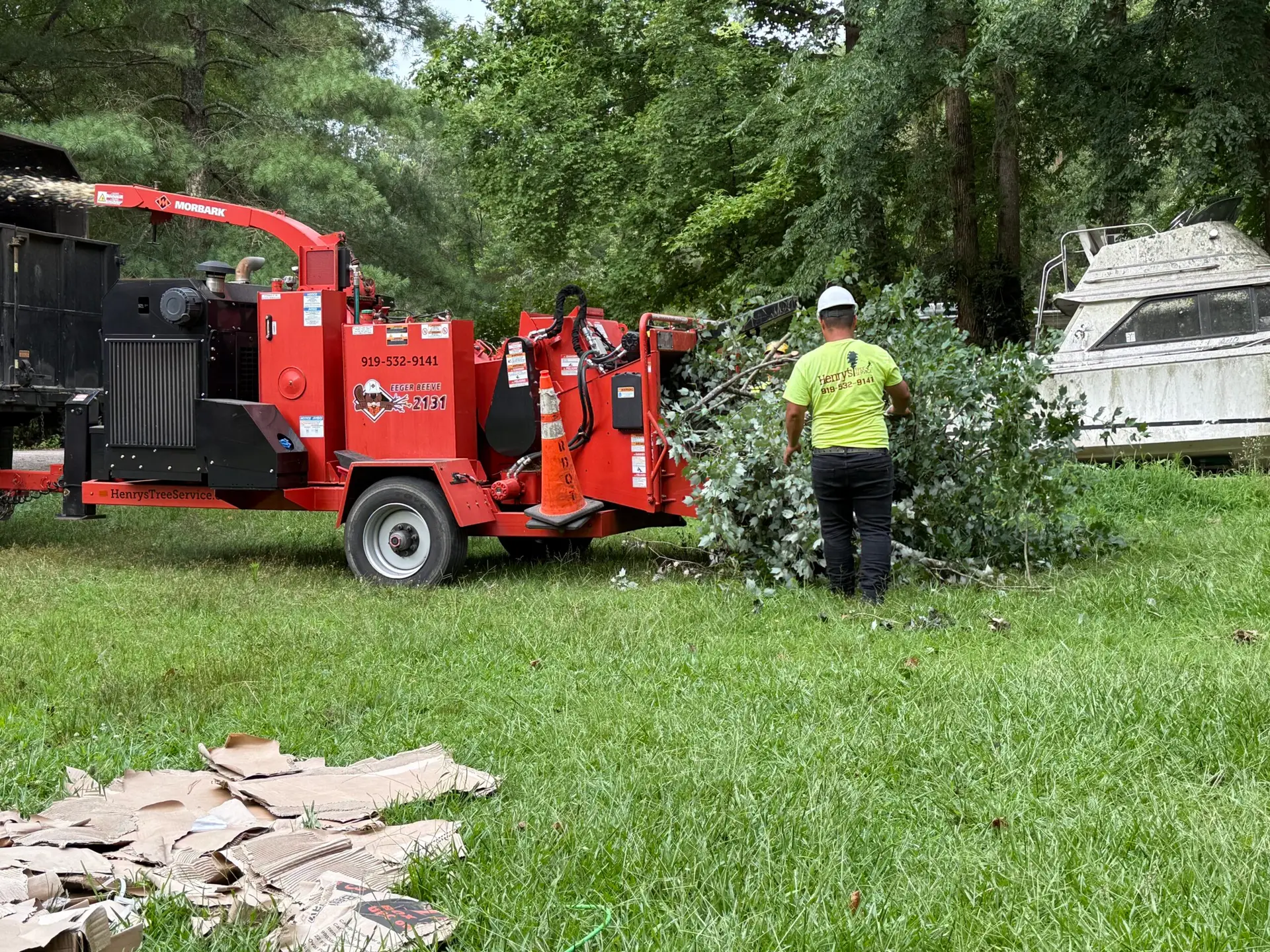 Tree care professional operating a chipper machine at a residential property, with fallen branches and cardboard on the ground, showcasing Henry's Tree Service equipment and expertise in tree removal and maintenance.